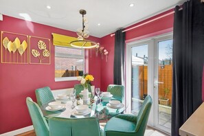 Een eethoek met een glazen tafel, groene beklede stoelen en een gele hanglamp.