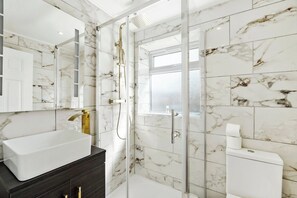 Een badkamer met een wit wastafel, donkerder badkamerkastje, marmeren wanden en een douche met glazen wanden.