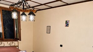 A room with two beds, one with a pink bedspread and the other with a blue bedspread. There is a window with a view of trees, a chandelier, and a framed picture on the wall.