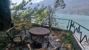 A rustic outdoor seating area with a stone table and chairs, overlooking a body of water and mountains.