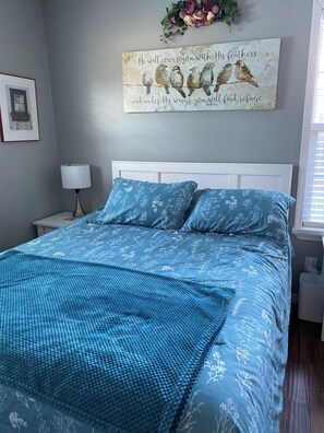 Une chambre à coucher comprenant un lit, une table de chevet, une lampe, un élément de décoration murale et une fenêtre.