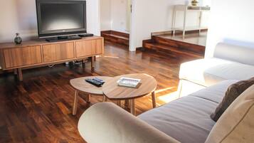 Un soggiorno con una TV a schermo piatto su un mobile in legno, due tavolini con telecomandi, un divano e una scala che conduce in un'altra stanza.