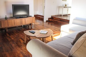 Een woonkamer met een flatscreen televisie op een houten kast, twee ronde tafels met afstandsbedieningen, een bank en een trap naar een andere kamer.