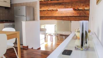 Una cucina moderna con frigorifero integrato, travi in legno a soffitto, zona pranzo con tavolo e sedie, e angolo living con divano e tavolino rotondo.