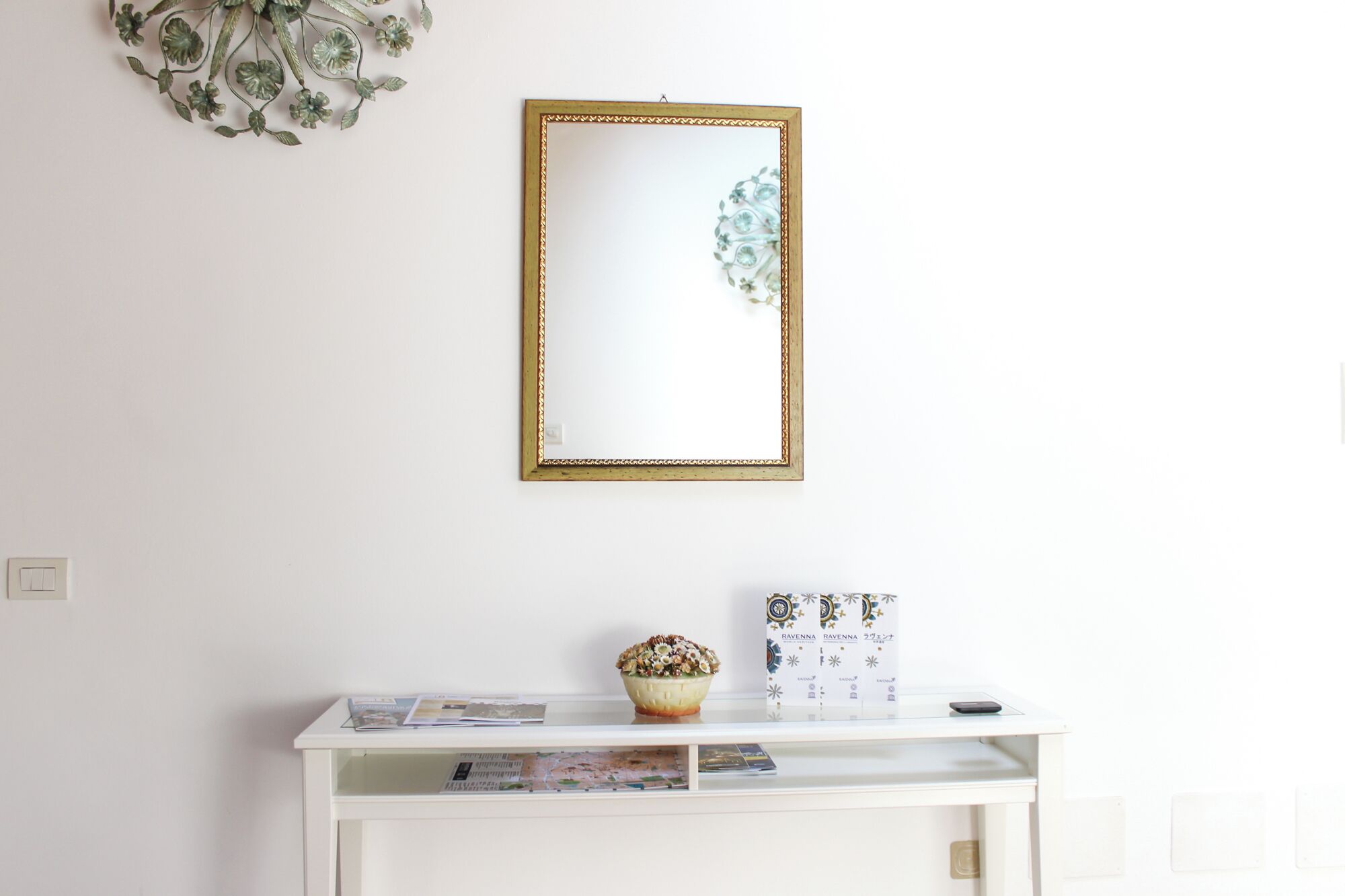 A white console table with a mirror, a bowl of flowers, and a box of magazines.
