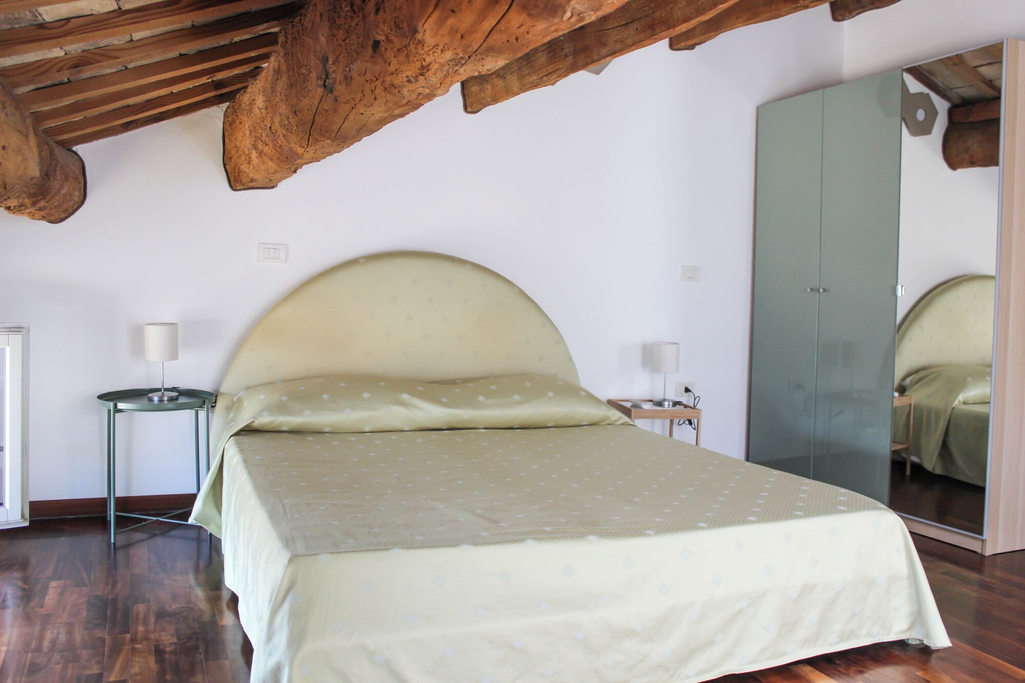 Une chambre à coucher avec un grand lit, des poutres apparentes en bois au plafond, un miroir et une petite table de chevet avec une lampe.