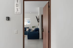 A doorway leading to a living room with a blue sofa, a wall-mounted light fixture, and a framed picture. - Ground Floor · 2 Pools · Balcony · 15 min to Beach (Mezcales)