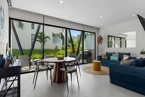 A modern living room with a large glass wall, a round dining table, and a blue sofa. - Ground Floor · 2 Pools · Balcony · 15 min to Beach (Mezcales)