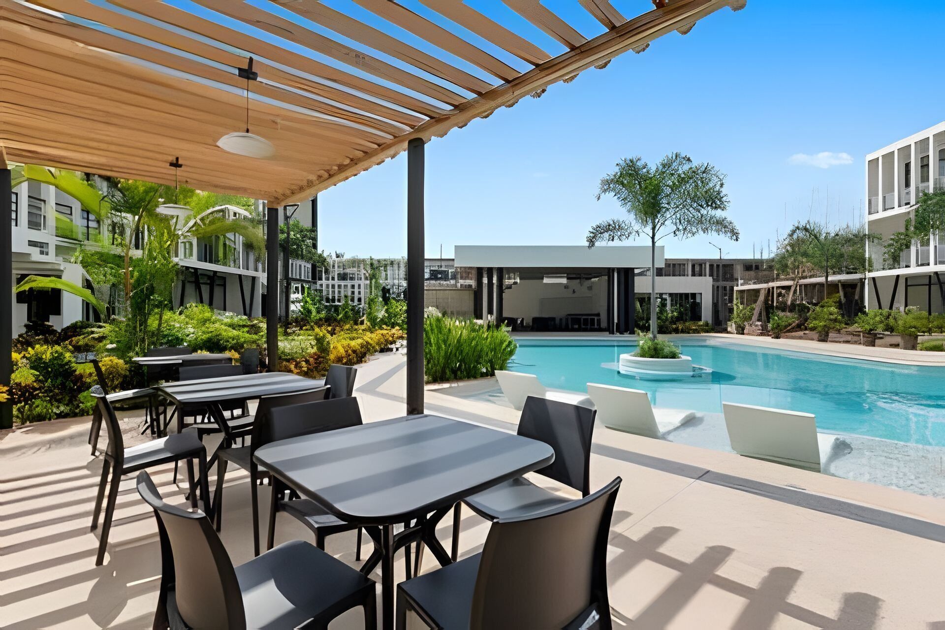 An outdoor pool area with tables and chairs, surrounded by greenery and modern buildings.