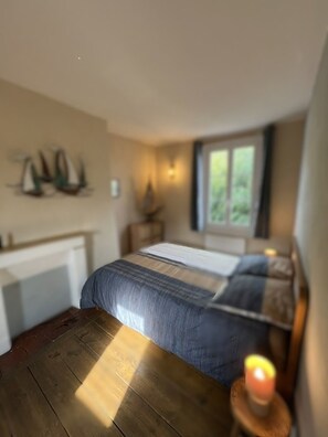 A bedroom with a wooden floor, a bed with a striped bedspread, a nightstand with a lamp, and a window with a view of greenery. - The Blue House in the Heart of St. Valéry-sur-Somme (St Valéry sur Somme)