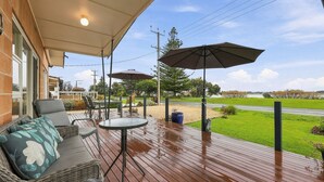 Un patio exterior con terraza de madera, zona de estar, sombrillas y vistas a una carretera y vegetación.