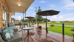 An outdoor patio with a wooden deck, seating area, umbrellas, and a view of a road and greenery. - 25 Captain Sturt Rd, Hindmarsh Island (Hindmarsh Island)