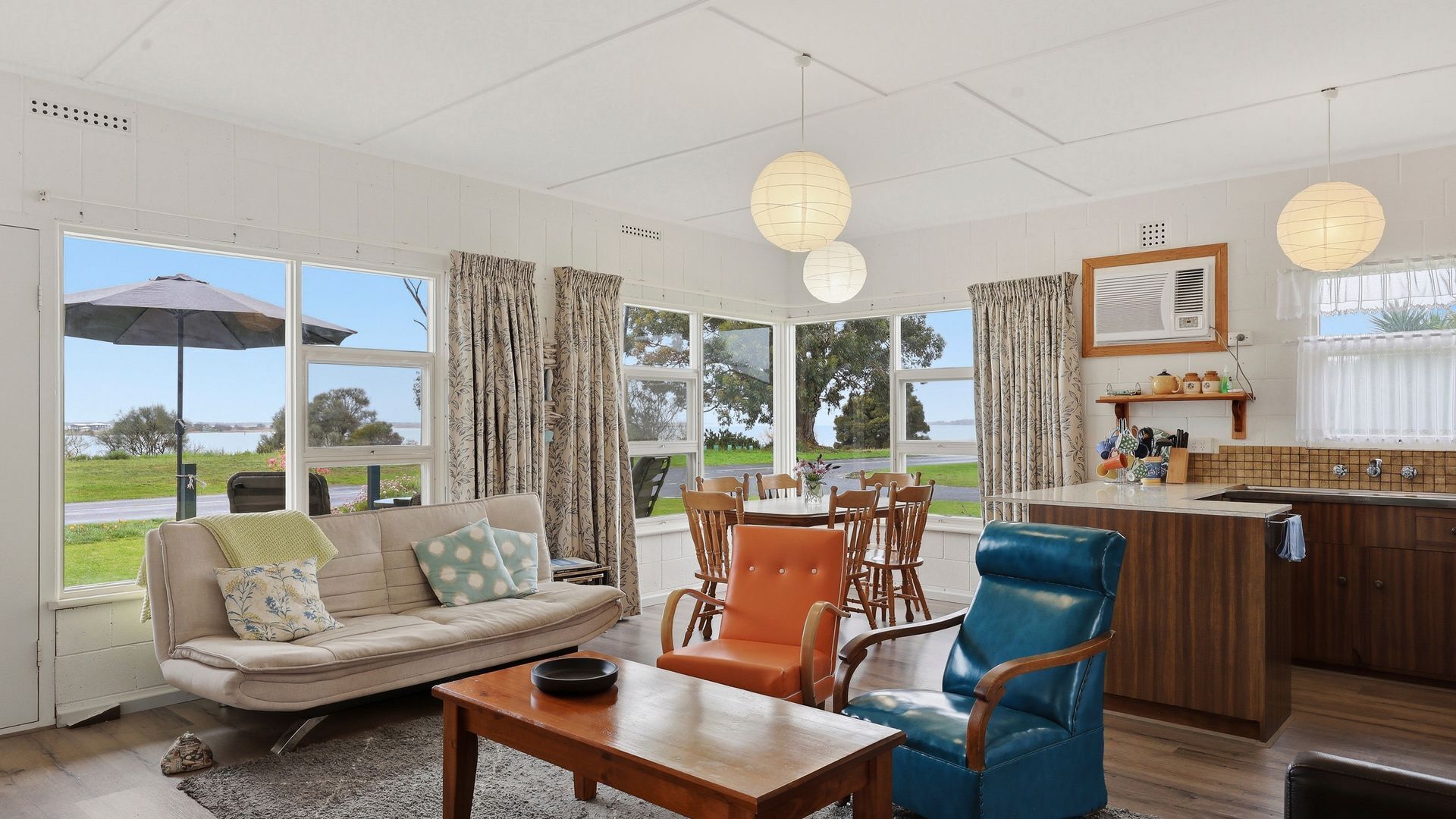 A spacious living area with a sofa, dining table, and chairs, large windows offering a view of the outdoors, and a kitchen area with a sink and counter.
