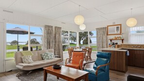 A spacious living area with a sofa, dining table, and chairs, large windows offering a view of the outdoors, and a kitchen area with a sink and counter. - 25 Captain Sturt Rd, Hindmarsh Island (Hindmarsh Island)