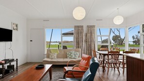 A spacious living room with a large window, a sofa, a dining table with chairs, and a flat-screen TV mounted on the wall. - 25 Captain Sturt Rd, Hindmarsh Island (Hindmarsh Island)