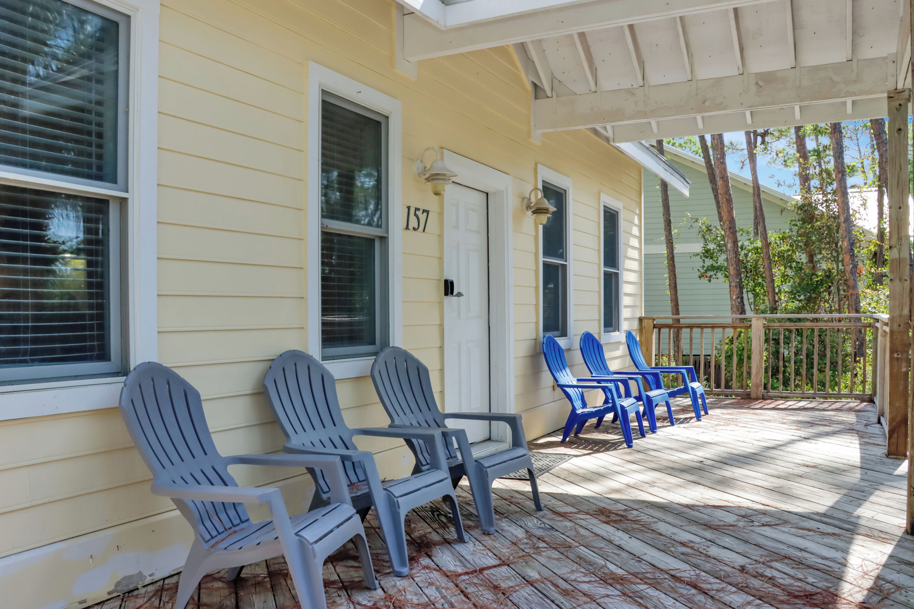 A porch with two grey Adirondack chairs and two blue Adirondack chairs, a white door with the number 157, and a wooden deck.