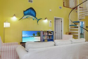 A room with a television displaying a beach scene, a bookshelf, a staircase, and a wall-mounted decorative piece. - Sunny Daze | Steps to Beach | Community Pool | Pet Friendly (Indian Pass)