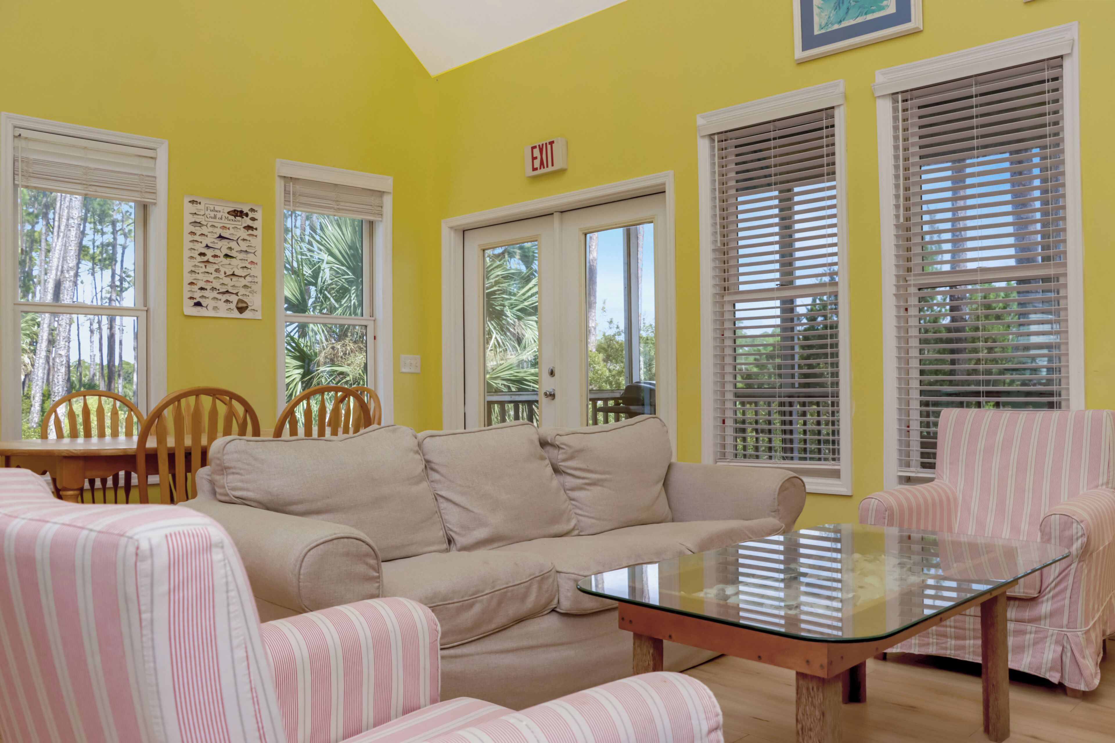 Una habitación con un sofá color beige, sillones a rayas, una mesa de centro de cristal y una mesa de comedor con sillas. Hay ventanas con persianas, una puerta y una señal de salida en la pared.