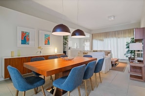A modern dining area with a wooden table and blue chairs, a kitchen in the background, and a living area with a sofa and curtains. - 5BR home with 5 baths at Windsor Hills (Kissimmee)