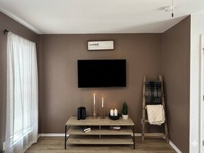 A living room with a flat-screen TV mounted on the wall, a wooden coffee table with candles, a ladder, and a small potted plant. - Modern 1BR Near AT&T Stadium + Six Flags (Arlington)