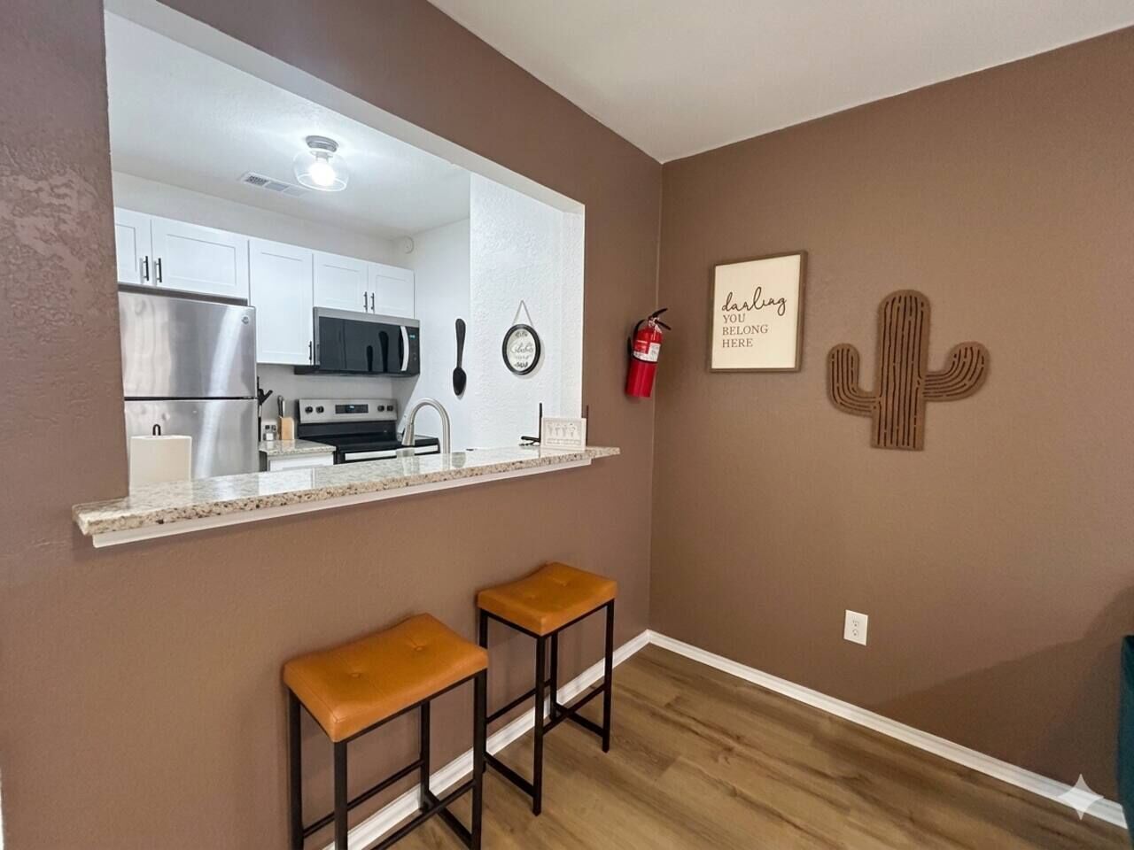 A kitchen with white cabinets, a stainless steel refrigerator, and a countertop. There are two stools and a fire extinguisher mounted on the wall.