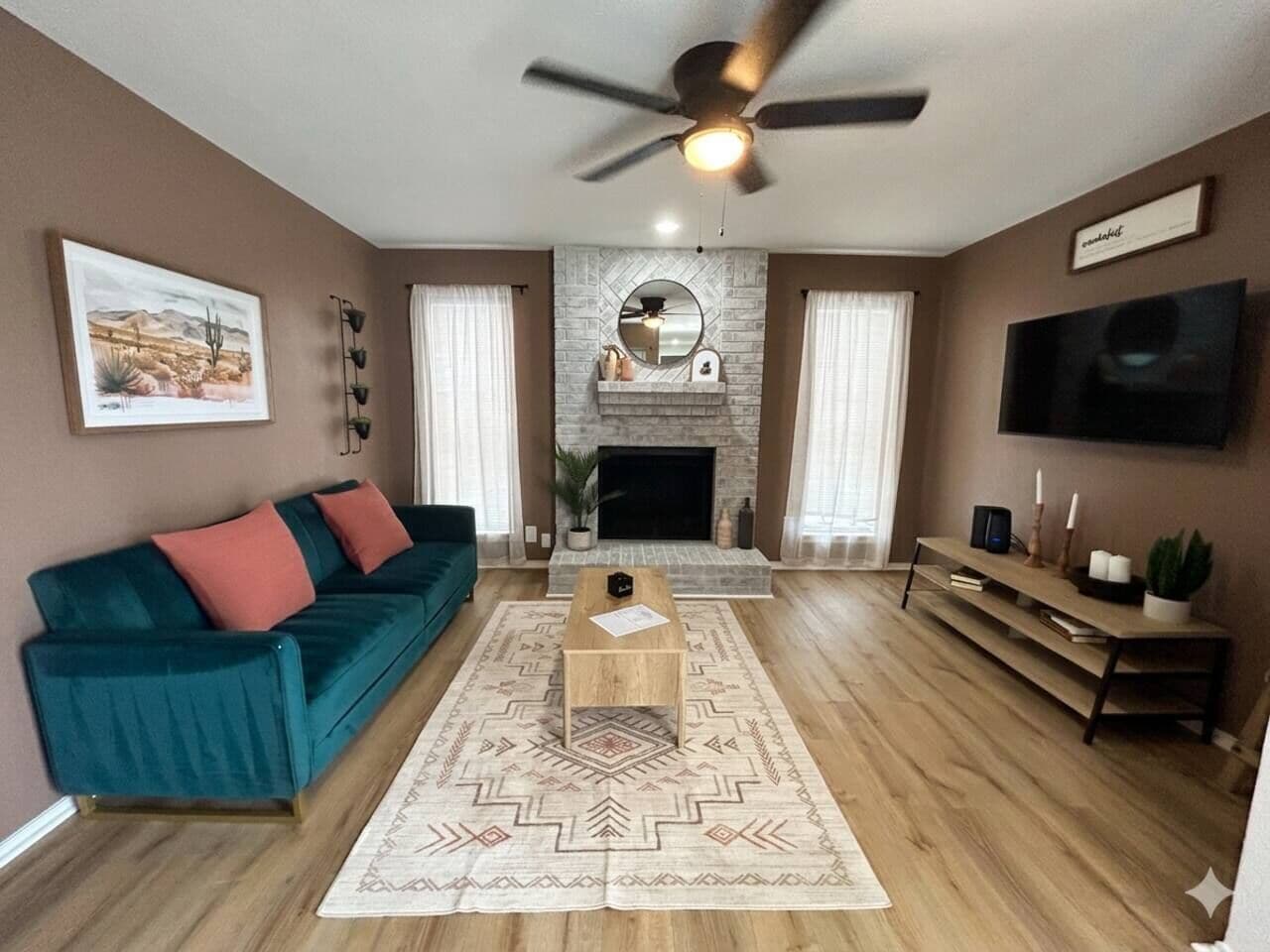 A living room with a teal sofa, a wooden coffee table, a fireplace, a ceiling fan, and a TV mounted on the wall.