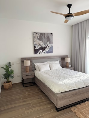 A modern bedroom with a wooden bed frame, white bedding, a ceiling fan, a wall-mounted artwork, a bedside lamp, and a potted plant. - Isla Azul Pool & Golf Views, 5 Min to Beach, Near Hard Rock (Punta Cana)