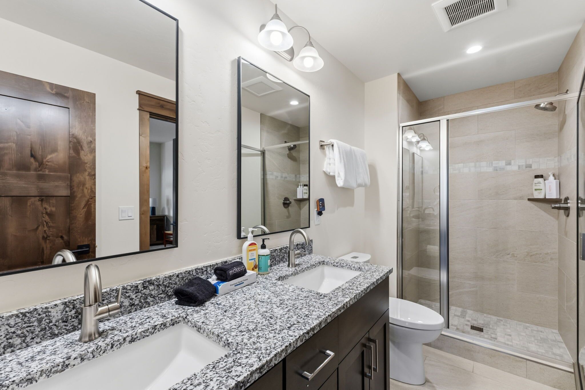 Un baño moderno con encimera de granito, lavamanos dobles, un espejo grande y una cabina de ducha de vidrio.