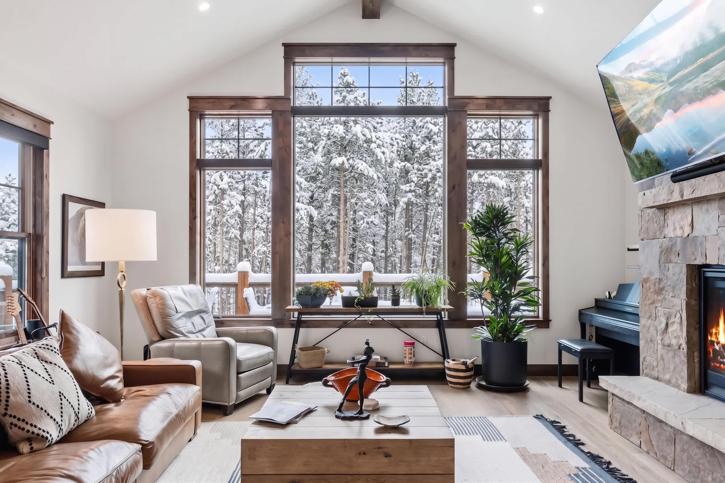 A cozy living room with a fireplace, a piano, and a large window overlooking a snowy forest.