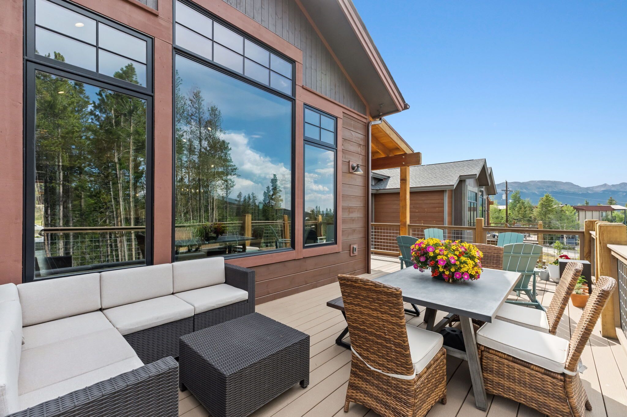 Une terrasse dotée de mobilier d’extérieur, d’une table agrémentée d’un arrangement floral et de grandes fenêtres donnant sur une zone boisée.