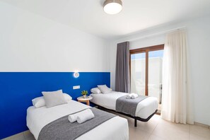 A hotel room with two beds, a small table, a nightstand, a window with curtains, and a view of the outside. - Villa Naira by Villa Plus (Playa Blanca)