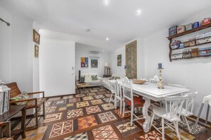 Un coin repas avec une table et des chaises blanches, un sol à motifs et une étagère remplie de conserves.