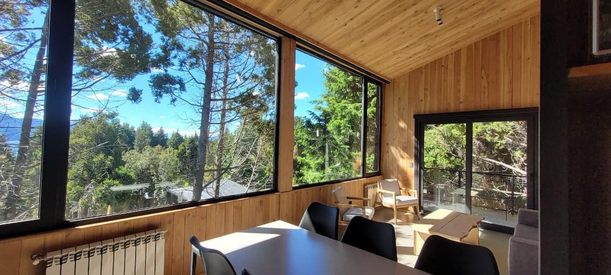 A room with a large window offering a view of trees and mountains. There is a table with chairs and a radiator below the window.
