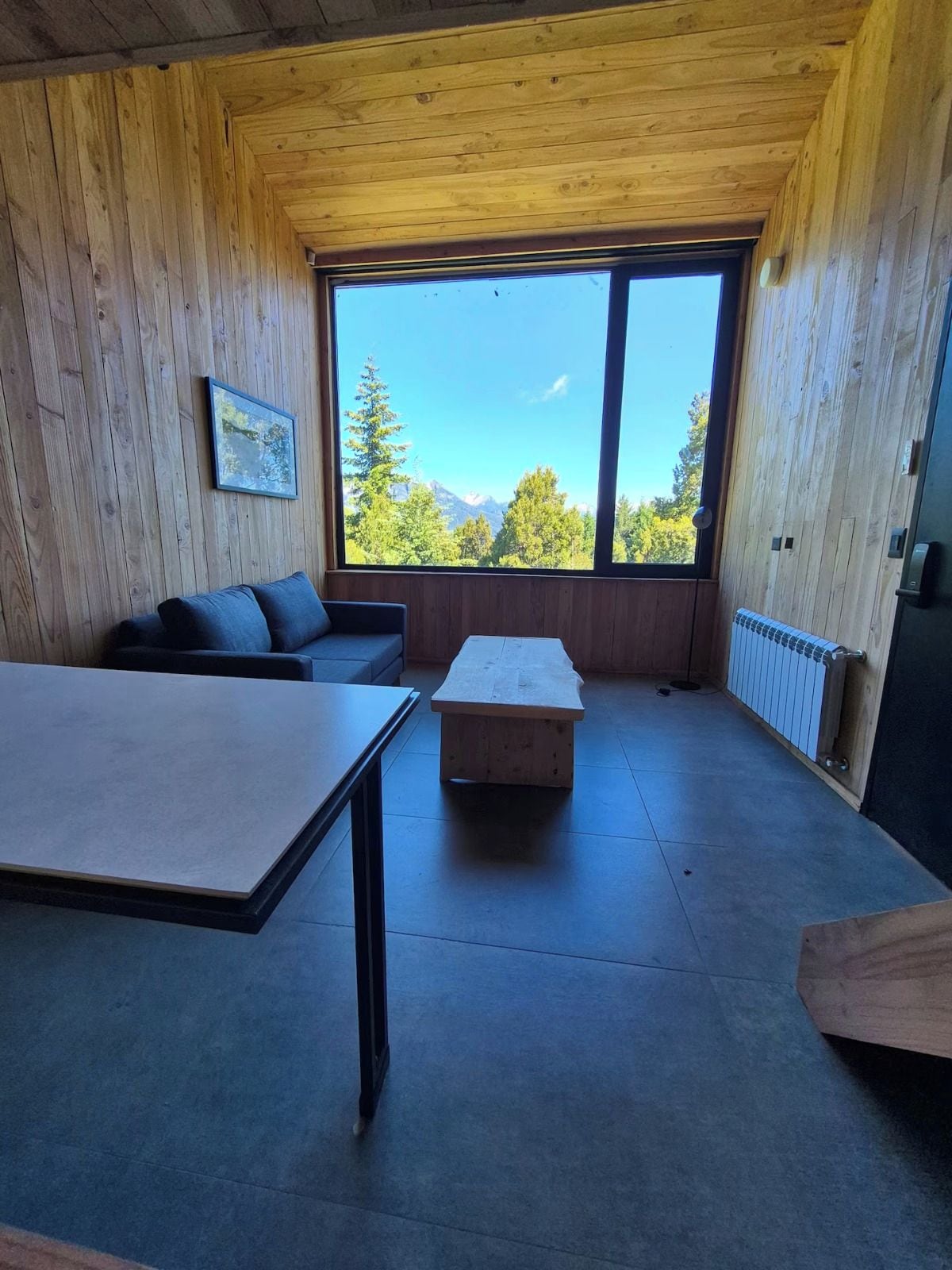 A room with wooden walls, a large window with a mountain view, a sofa, a table, and a radiator.