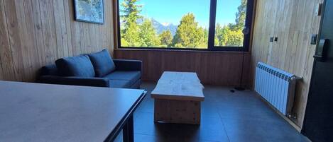 A room with wooden walls, a large window with a mountain view, a sofa, a table, and a radiator.