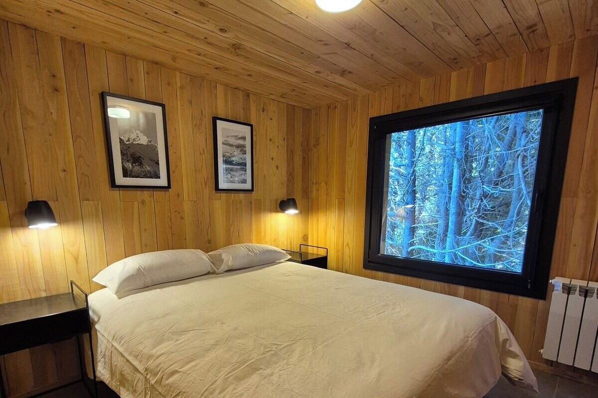 Un dormitorio con una cama, dos mesitas de noche, un ventanal con vistas a árboles y paredes de madera.