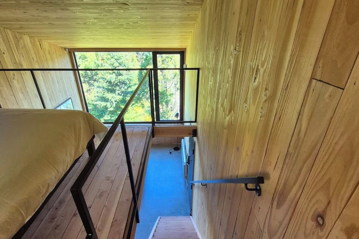 Un interior de madera con una cama litera, una escalera y un ventanal con vistas a la vegetación.