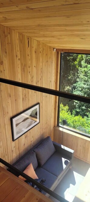 Una habitación con techo de madera, un ventanal grande, un cuadro enmarcado y un sofá oscuro con cojines.