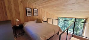 Dormitorio de cabaña de madera con cama, mesita de noche, lámpara y ventana con vistas a un bosque.