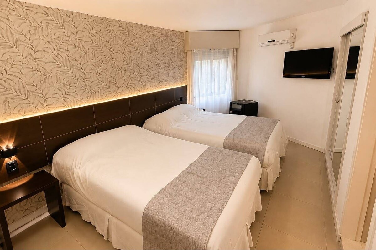 A hotel room with two beds, a nightstand, a TV, and a window with curtains.