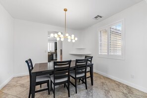 A dining area with a rectangular table, four chairs, a chandelier, and a window with blinds. - Lovely 5-bedroom 3 Full Bathroom House In Charming Southwest Bakersfield (Bakersfield)