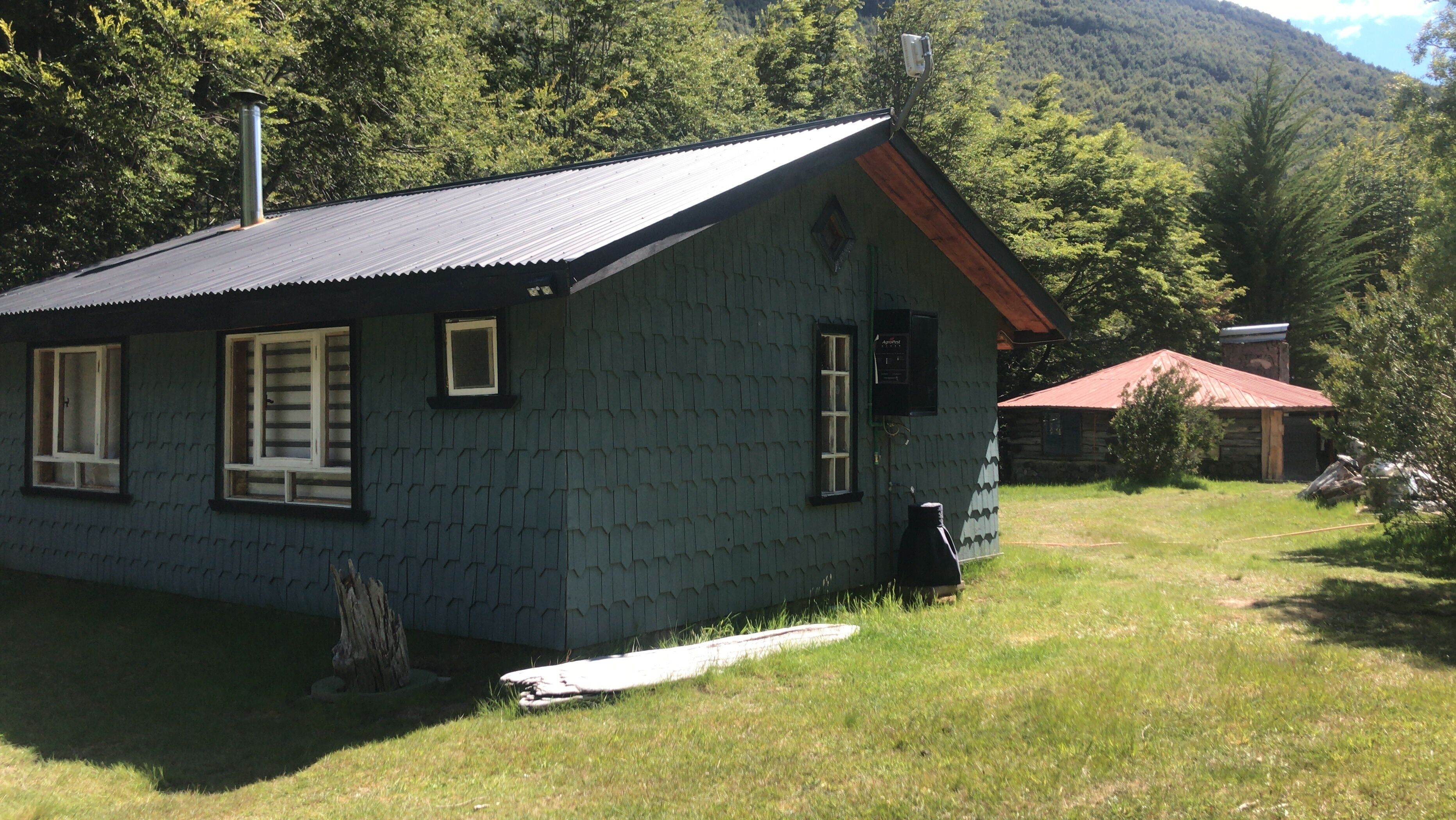 A small, single-story cabin with a sloped roof, surrounded by a grassy area and dense forest.