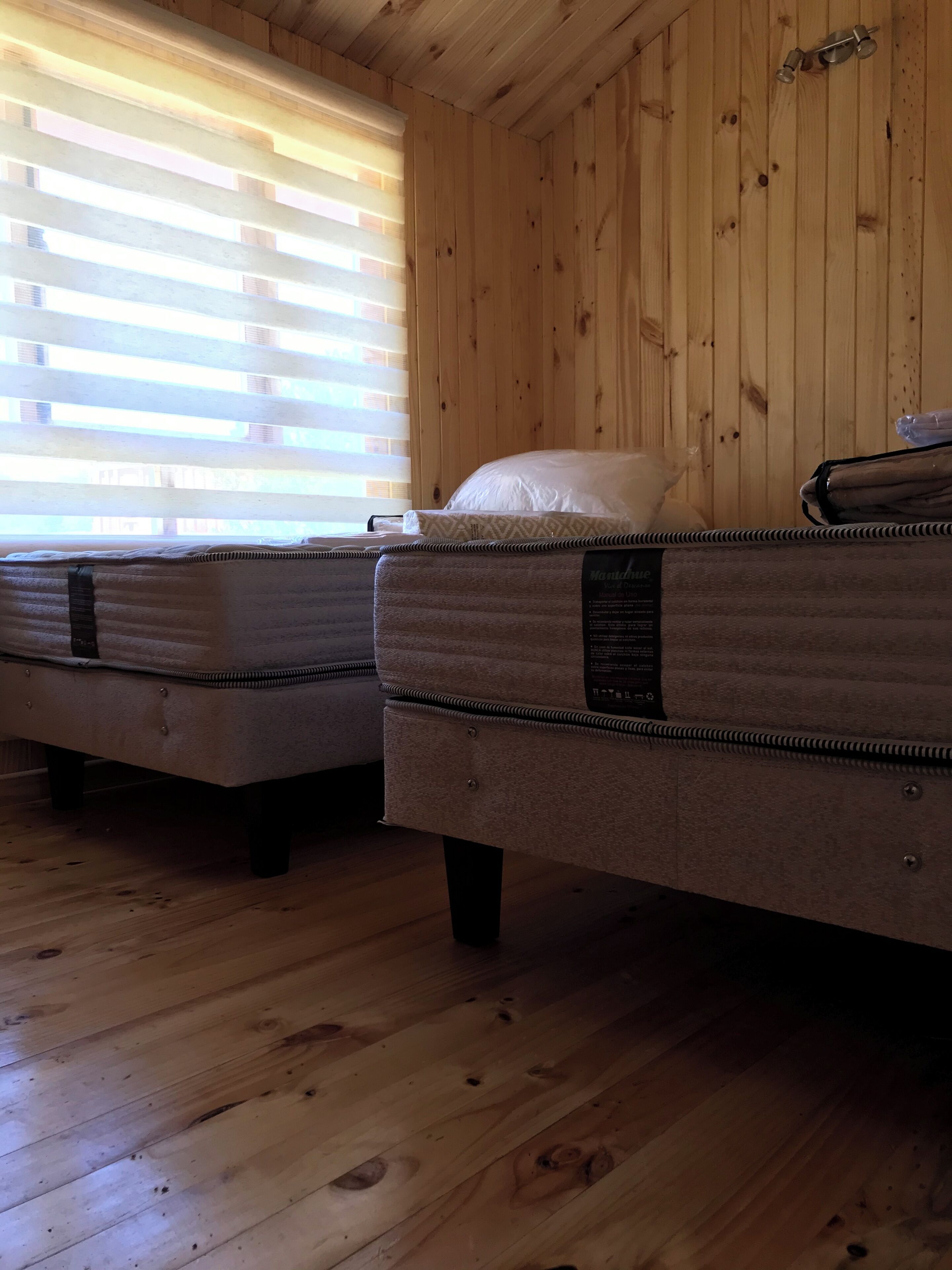 A room with wooden walls and floors, two beds with mattresses and pillows, and a window with blinds.