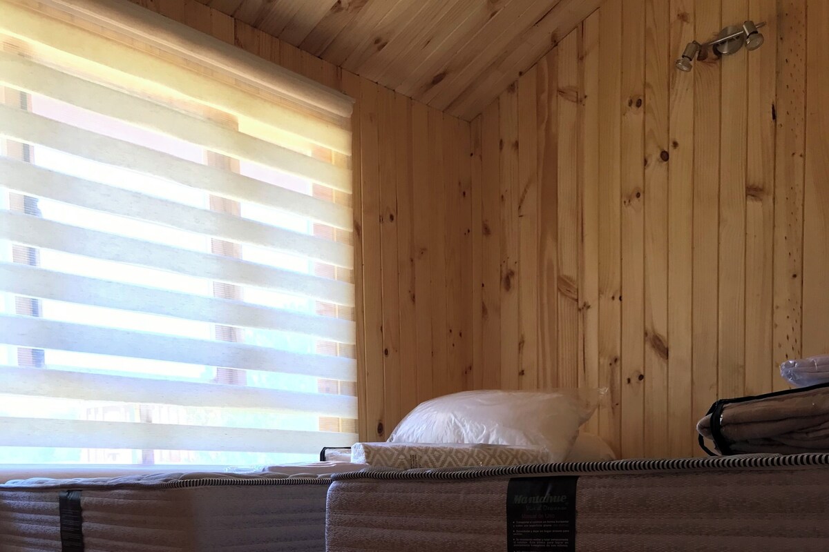 A room with wooden walls and floors, two beds with mattresses and pillows, and a window with blinds.