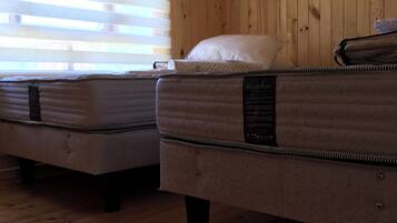 A room with wooden walls and floors, two beds with mattresses and pillows, and a window with blinds.