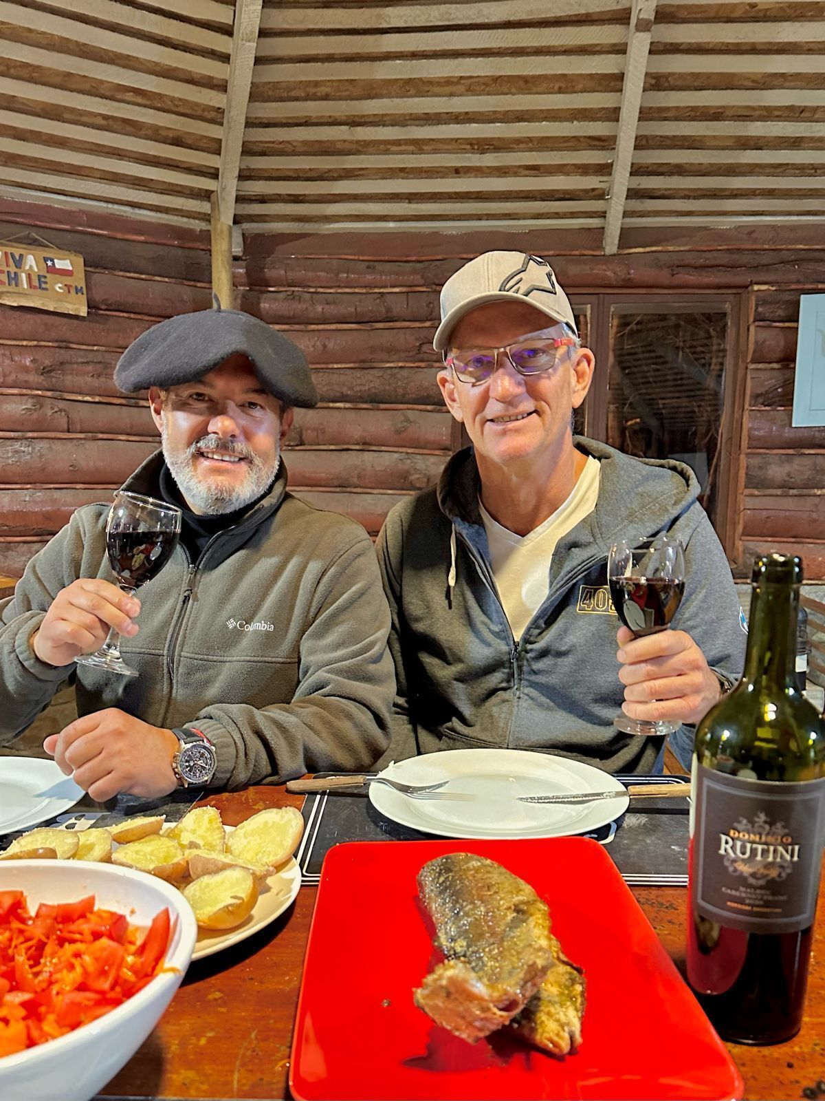 Two people seated at a wooden table with a red plate of food, a bottle of wine, and a bowl of vegetables.
