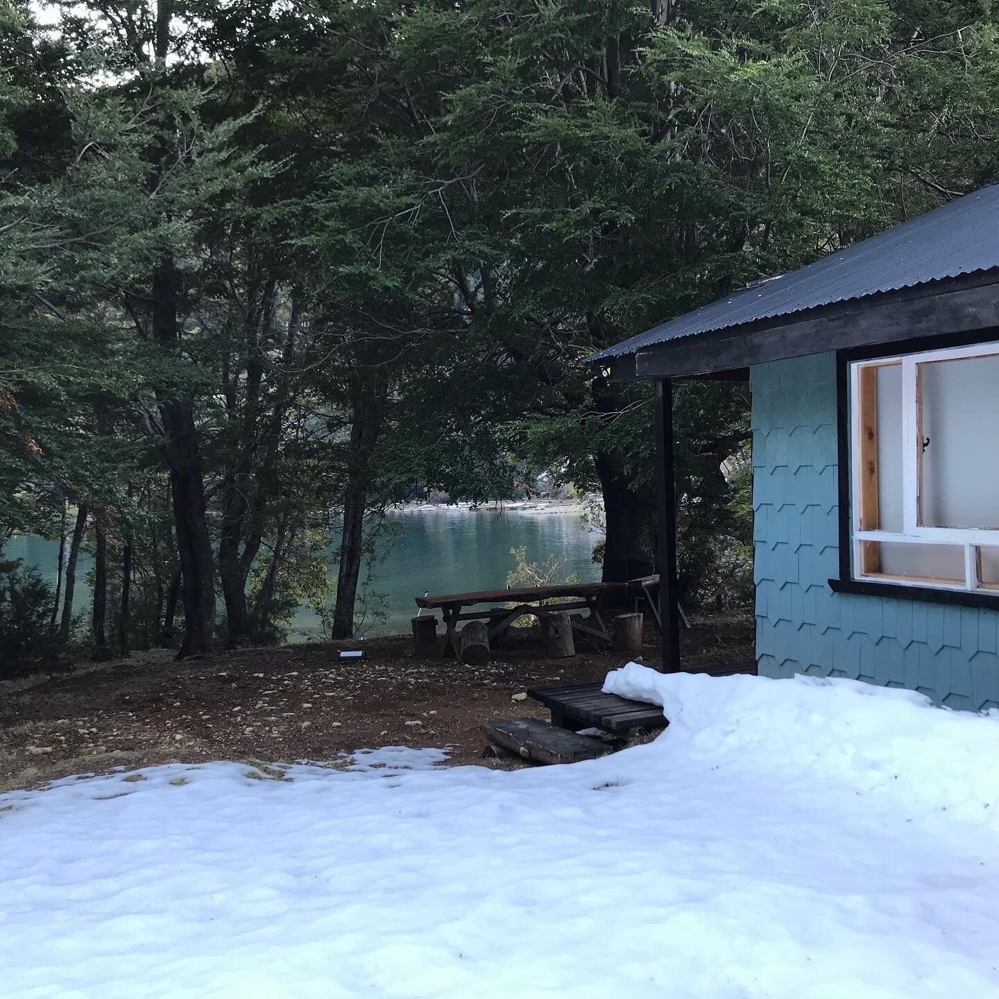 Une cabane dotée d’une terrasse en bois et d’un banc, entourée d’arbres et bordée par un plan d’eau.