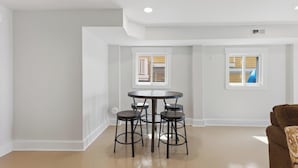 Um interior moderno com uma mesa redonda e quatro banquetas, uma janela com vista para um edifício, e um sofá castanho.
