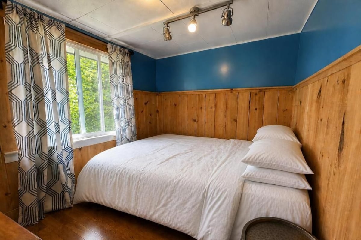 A bedroom with a wooden headboard, a bed with white bedding, a window with patterned curtains, and a wooden floor.