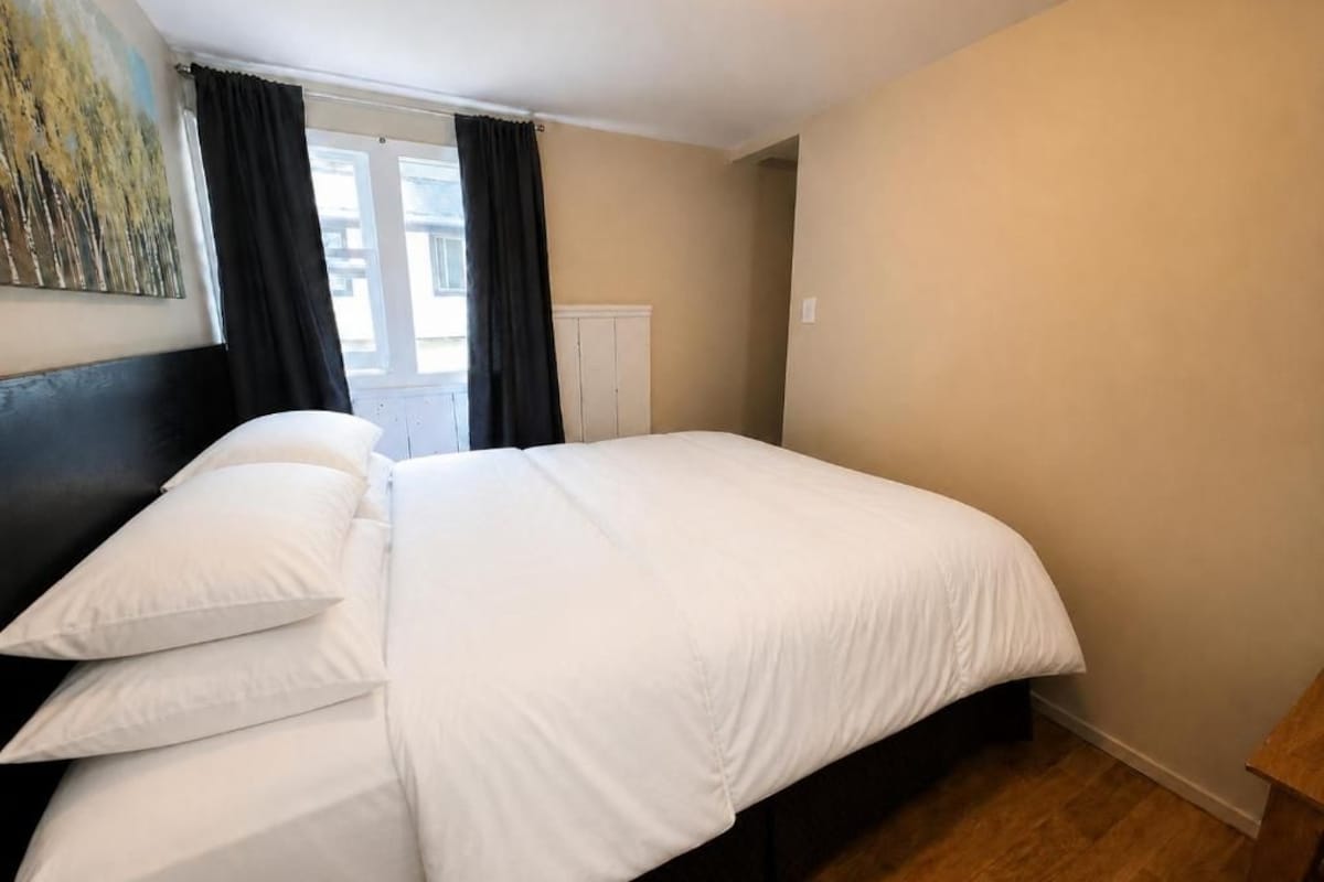 Une chambre à coucher avec un lit, une tête de lit, une fenêtre avec des rideaux et un tableau accroché au mur.